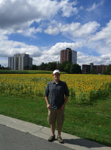 Pete at Farm with Civic in Background.jpg” with Caption: “Pete Anderson posing near the threatened experimental plots with the Civic Hospital in the background. Photo credit: Laura Cameron.