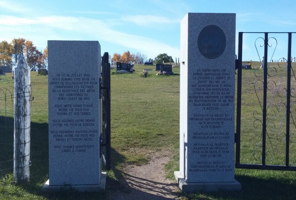 Two rectangular stone blocks frame the entrance to a cemetery. Both blocks have text inscribed on them. The shorter one on the viewer’s left has French text, while the one on the right has Michif text.
