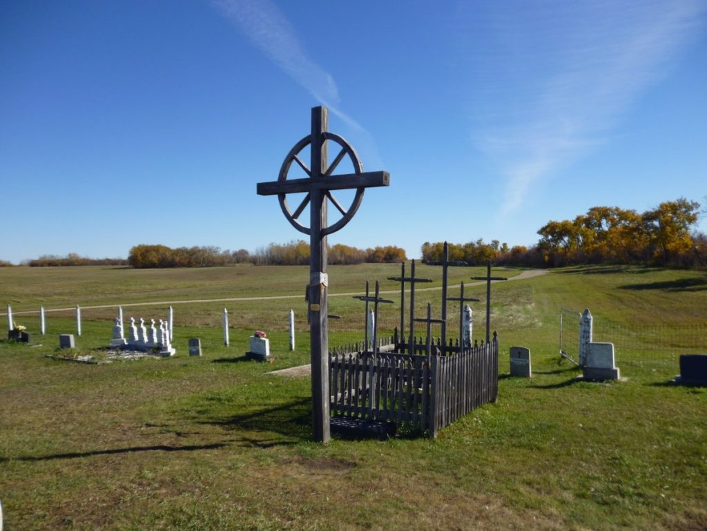A mass grave sits at the edge of a cemetery. It consists of a single large wooden cross with nine smaller wooden crosses standing behind it. The nine smaller crosses are surrounded by a short wooden fence.