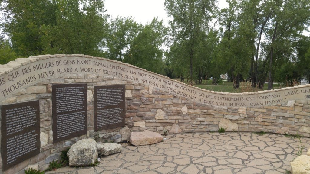 An image of a curving stone wall. The height of the wall goes up and down like a wave. Along the top of the wall is an inscription in both French and English. Embedded in the wall are three large plaques inscribed with text.