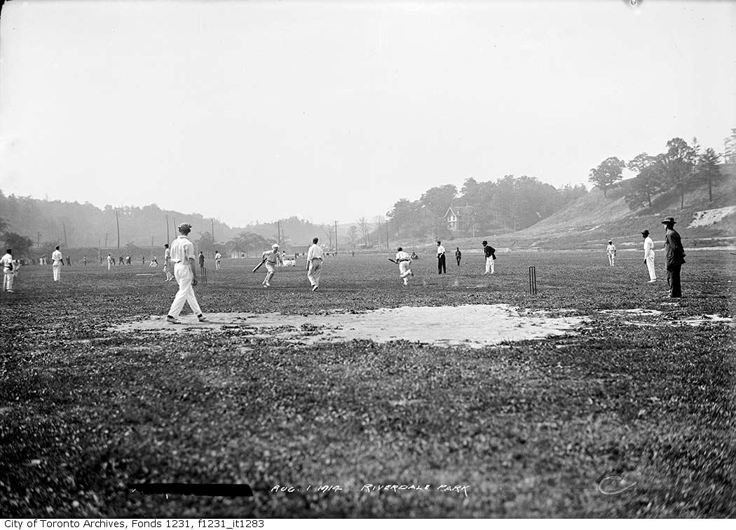Alt text: A black-and-white photo of several men playing cricket in a park. There is a treed hill in the background and a building is visible through the trees.