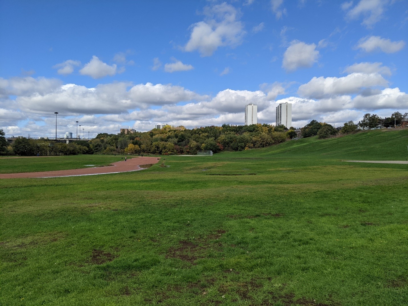 a colour photo of a lawn with a running track and baseball pitch. There is a hill on the left side of the picture and there are trees and high-rise buildings in the distance. The sky is blue with quite a few light grey clouds.