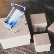 Four small brown boxes, one of which has a miniature shopping cart on top. There is a credit card in the shopping cart.