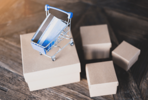 Four small brown boxes, one of which has a miniature shopping cart on top. There is a credit card in the shopping cart.
