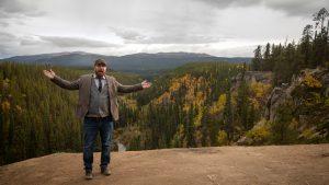 A man with a red beard stands in front of a treed vista with his arms outstretched.