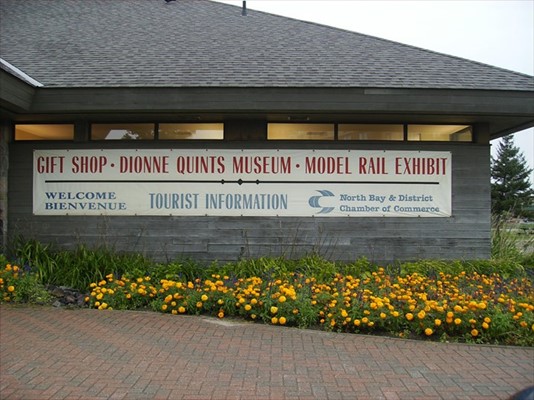 A single-storey wooden building with a large sign that reads "Gift Shop - Dionne Quints Museum - Model Rail Exhibit - Welcome Bienvenue - Tourist Information - North Bay & District Chamber of Commerce."