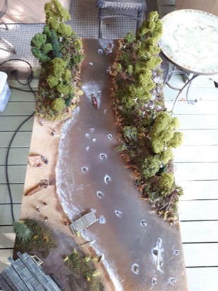 A birds-eye view of a miniature model representing a river with trees on the shore, two canoes, and a beach.