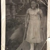 A smiling girl stands on a path, wearing a long white dress. She has one foot in a cast and is using crutches.