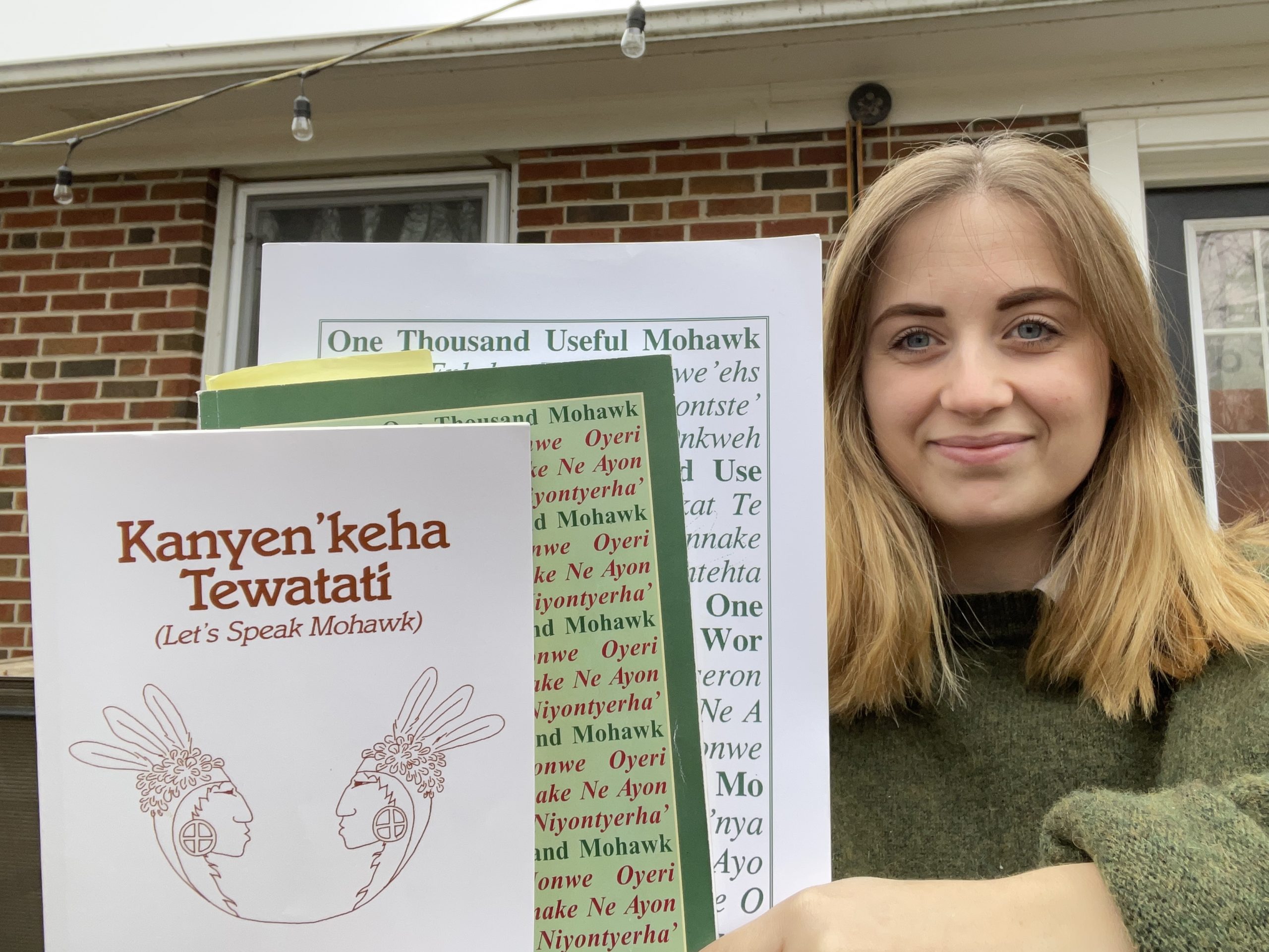 Photo of a blonde woman in a green sweater holding three books. The book at the front of the stack is titled "Kanyen'keha Tewatati (Let's Speak Mohawk)." 