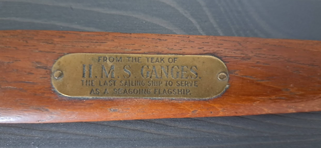 Close-up photograph of a brown piece of wood. There is a brass plate affixed with these words embossed on it: “From the teak of H.M.S. Ganges, the last sailing ship to serve as a seagoing flagship.”