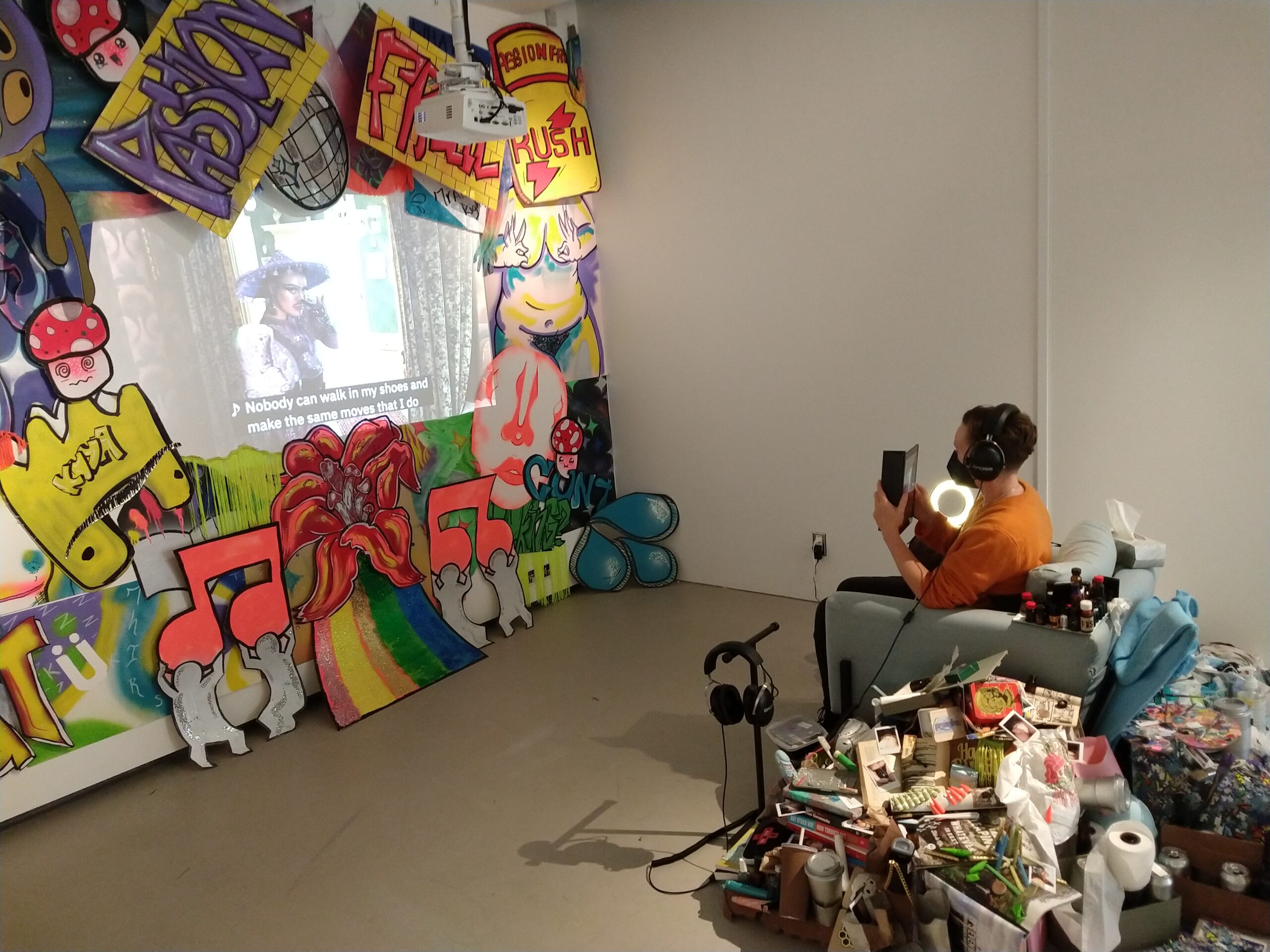 Photograph of a person in an orange shirt and black face mask wearing black headphones. They are looking at a video screen that is surrounded by brightly colour artwork. The person is surrounded with objects including a roll of toilet paper.