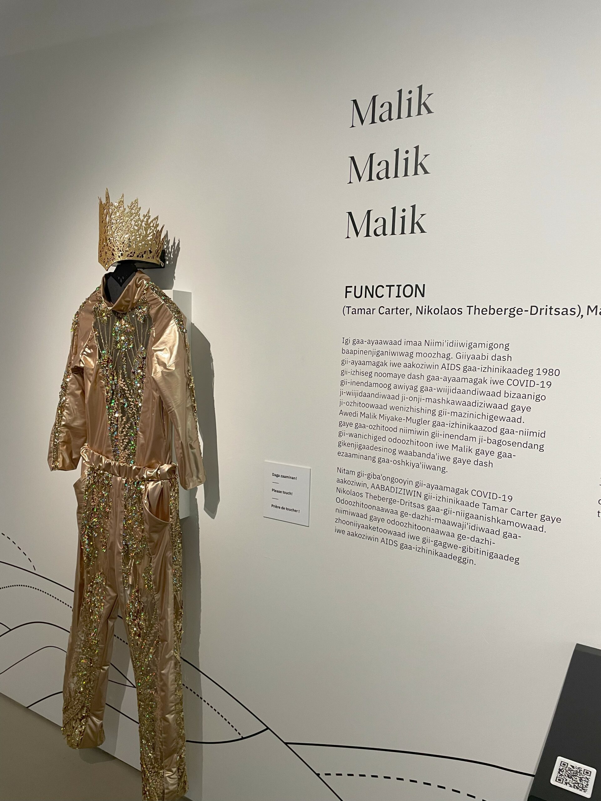 A white wall with black text. There is a gold bodysuit and headpiece hanging on the wall.