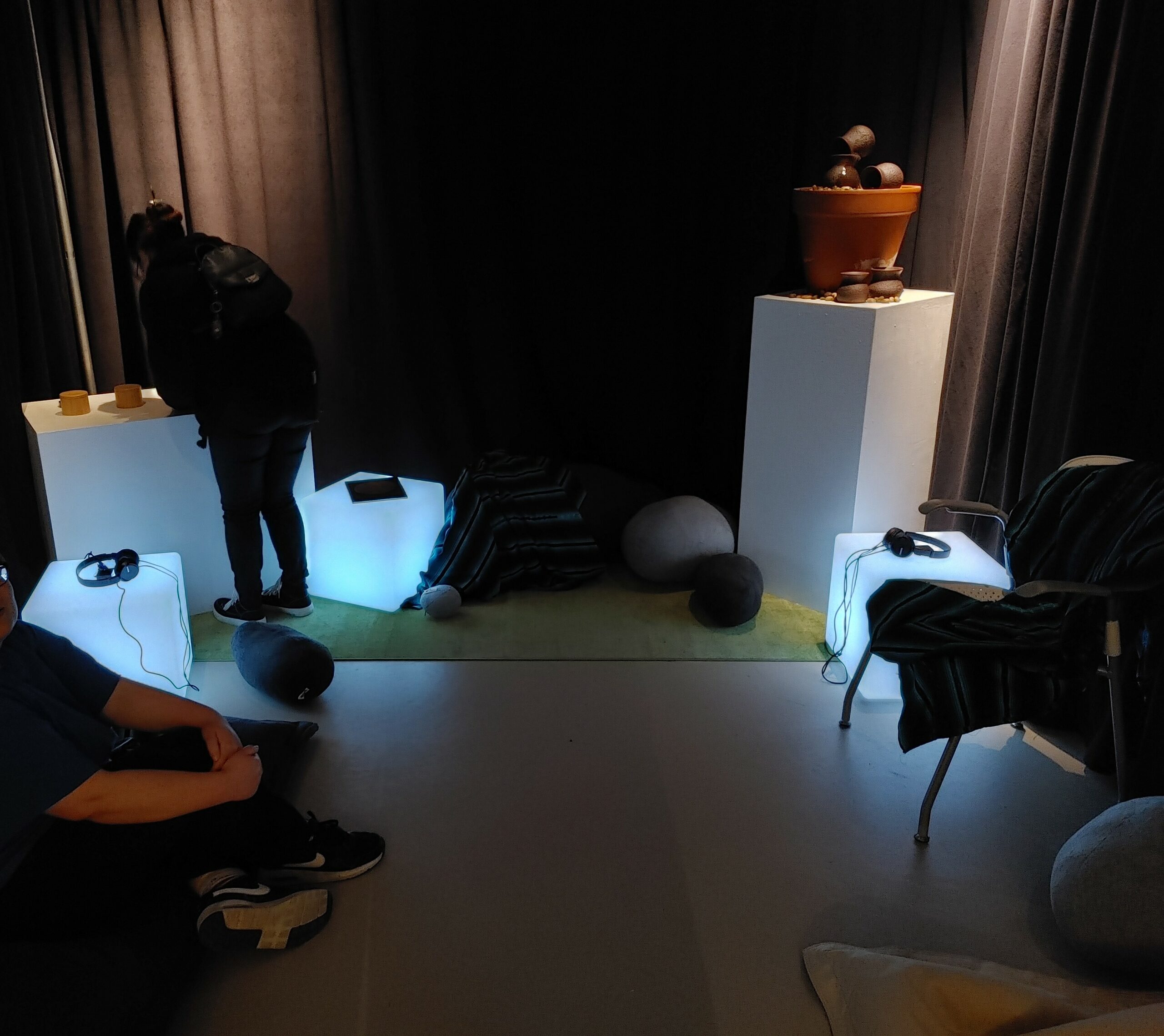 Two people in a darkened room, one sitting and one standing. There are softly glowing white boxes with headphones sitting on top.