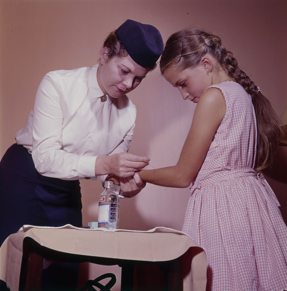 A nurse wearing a blue pillbox hat, white blouse, and dark blue skirt holds a young girl's arm. The nurse is using a needle and bottle of fluid to administer a skin test.
