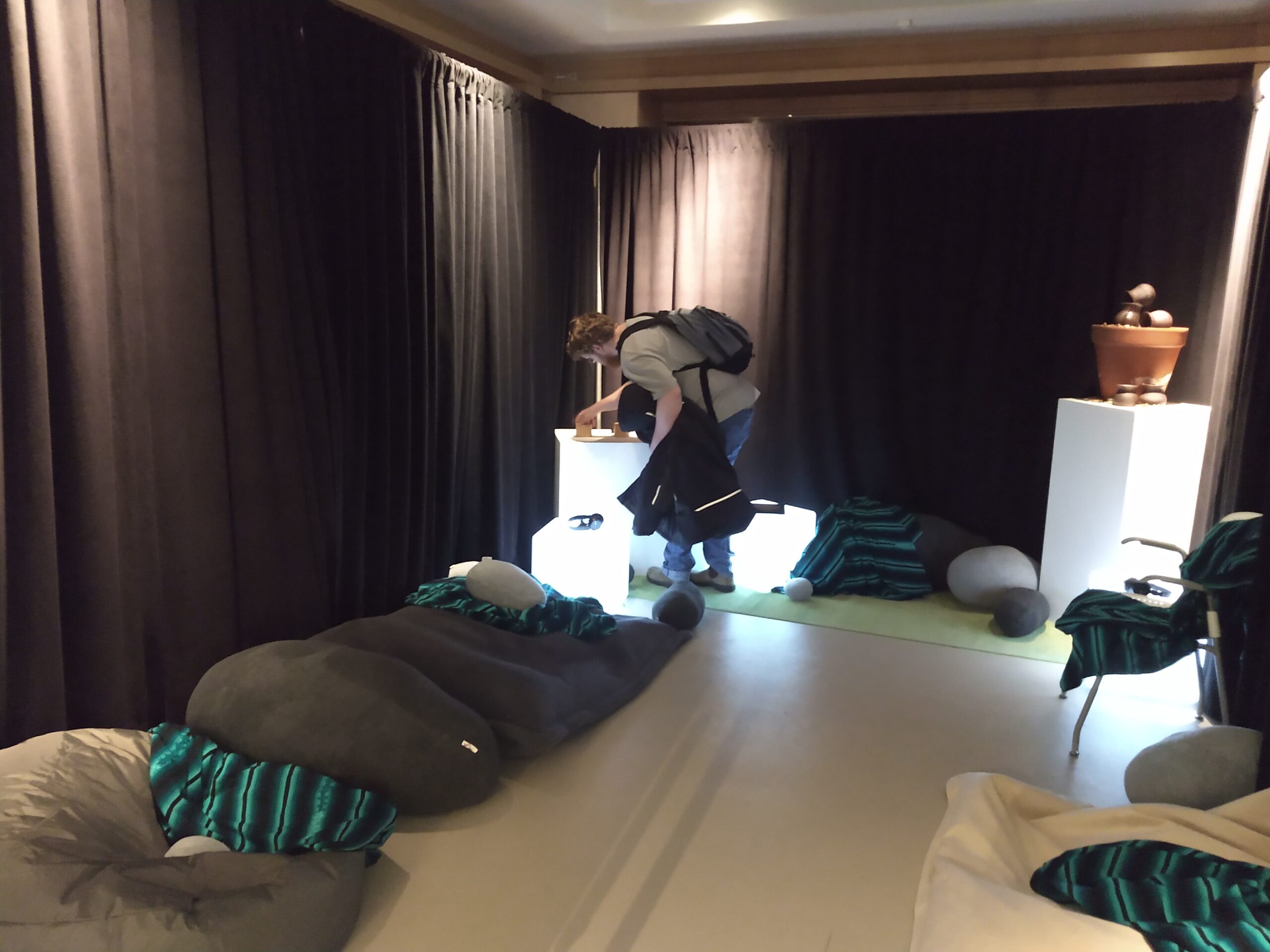 Photograph of a dimly lit room with black draped walls and soft cushions on the floor. There is a person in the corner wearing blue jeans, a beige t-shirt, and a black backpack.