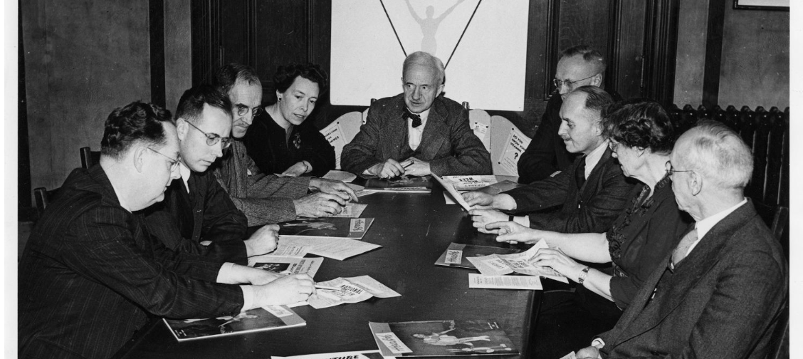 A black-and-white photograph of seven men and two women sitting around a conference table that is covered with papers and magazines. At the head of the table sits an older white man with a moustache.