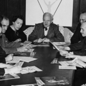 A black-and-white photograph of seven men and two women sitting around a conference table that is covered with papers and magazines. At the head of the table sits an older white man with a moustache.