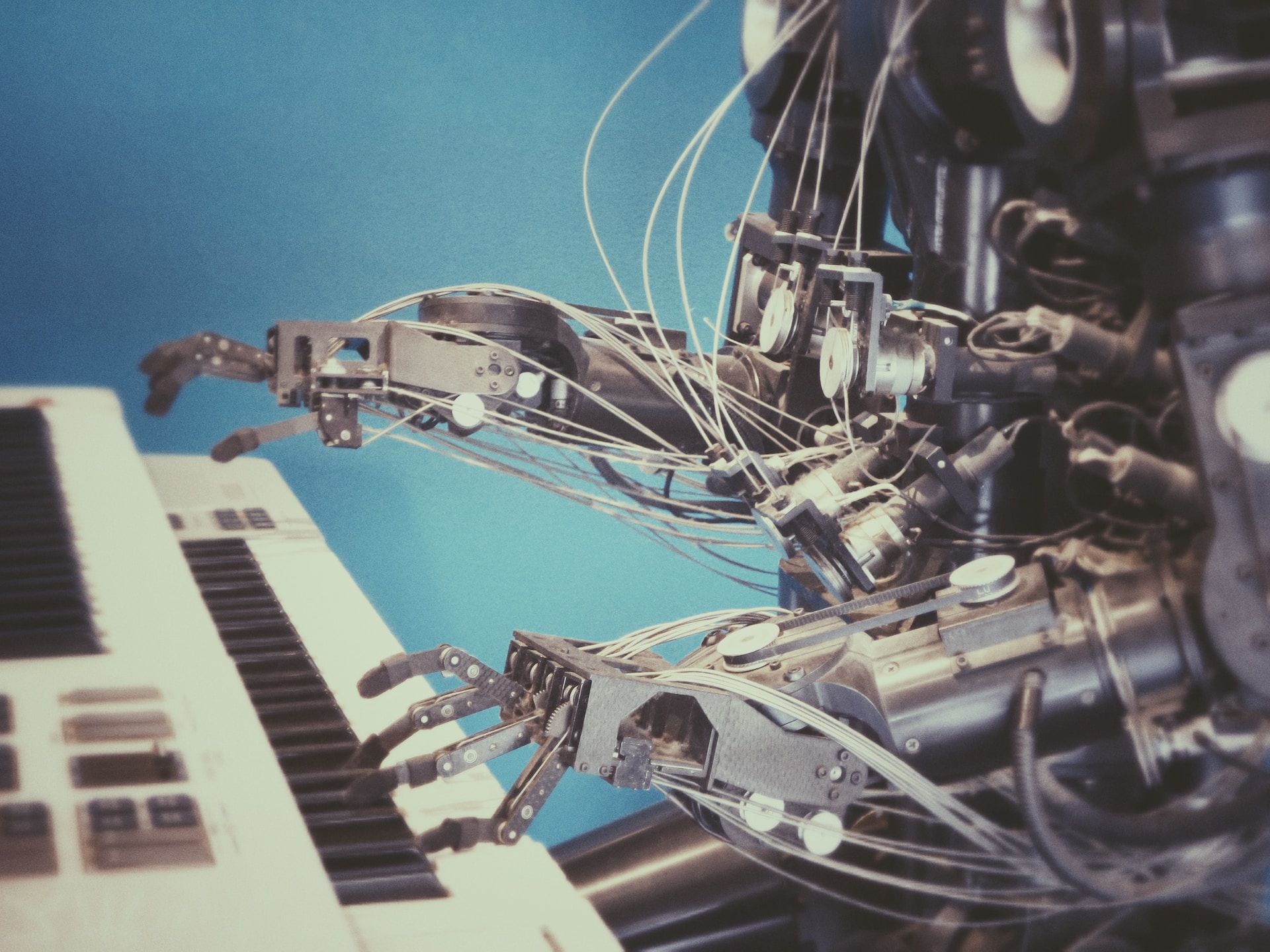 Colour photo of a metallic grey, humanoid robot with articulated hands playing a white piano keyboard with a blue background.