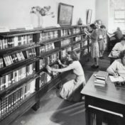 Black-and-white photograph of several women in a library, looking for books on the shelves and working at tables.