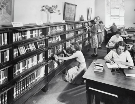 Black-and-white photograph of several women in a library, looking for books on the shelves and working at tables.