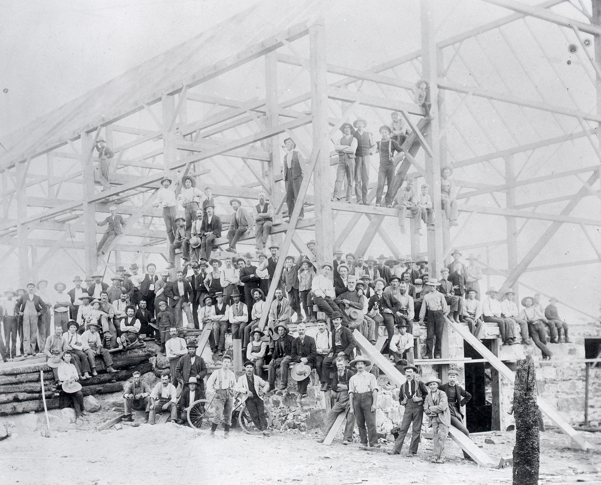 Black and white photo of many people posing around the frame of a barn.