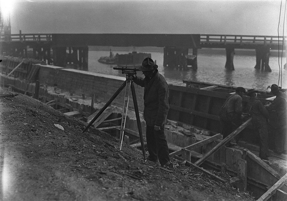 Black and white photo of a man on a construction site, sighting with a surveyor's level. There are three men behind him with their backs to the camera. A bridge spans a river in the background.
