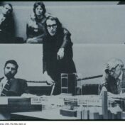 Photo of a man with long hair and glasses pointing at a building model. There are other people next to and behind the central figuure.