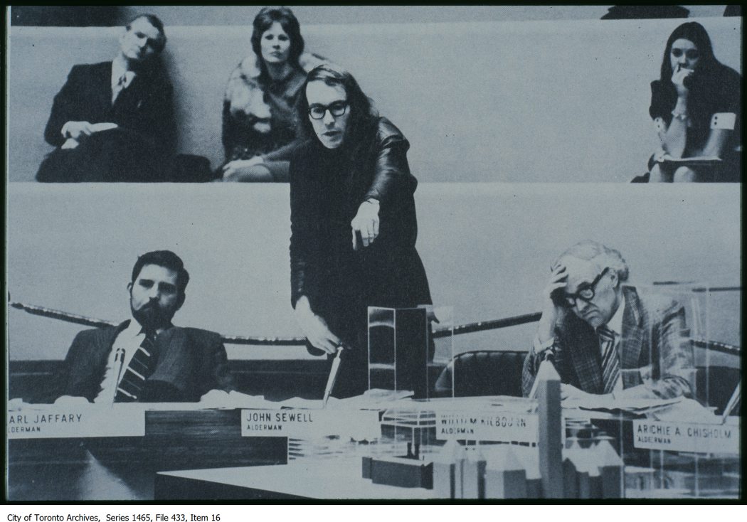 Photo of a man with long hair and glasses pointing at a building model. There are other people next to and behind the central figuure.