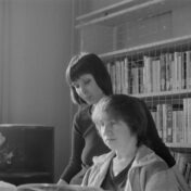 Black and white photo of two women looking at a book with bookshelves behind them.