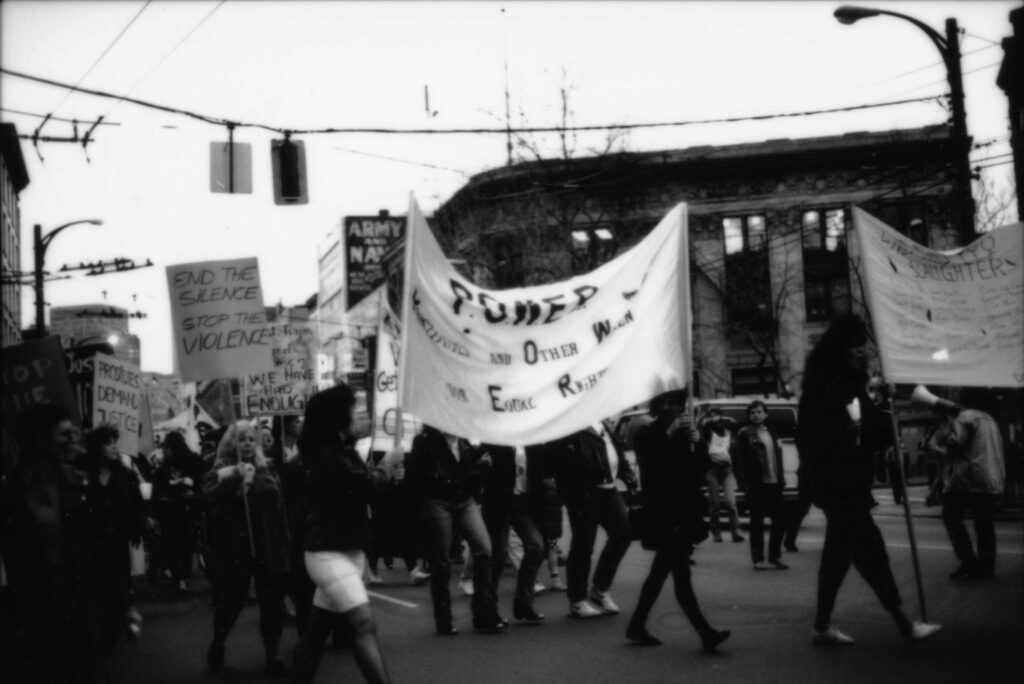 A black and white photo of a protest march on a city street. Marchers are carrying signs and banners with slogans like "POWER: Prostitutes and Other Women for Equal Rights", "End the Silence Stop The Violence", and "Prostitutes Demand Justice."