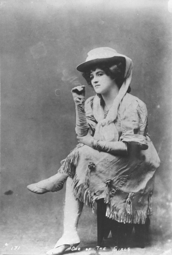 Black-and-white photo of a seated woman wearing a brimmed hat, fringed dress, and heeled boots.
