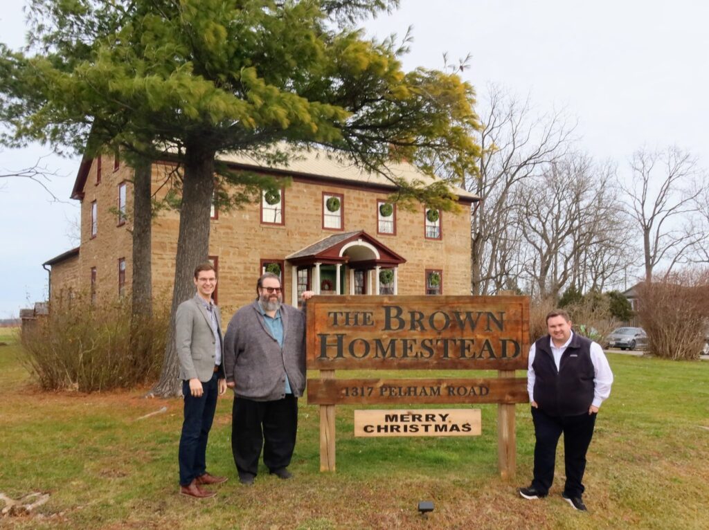 Three men standing in front of a large, brown stone historic home. They are standing around a sign that says "The Brown Homestead 1317 Pelham Road."
