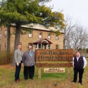 Three men standing in front of a large, brown stone historic home. They are standing around a sign that says "The Brown Homestead 1317 Pelham Road."
