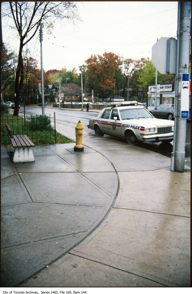 Colour photo of a city street corner with a police car marked "Metro Police" in the midground. Labeled "City of Toronto Archives, Series 1465, File 169, Item 144."