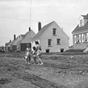 Black and white photo of children standing on a dirt road. Behind them, a line of detached homes extends into the distance.