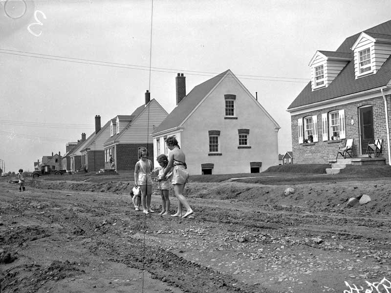 Black and white photo of children standing on a dirt road. Behind them, a line of detached homes extends into the distance.