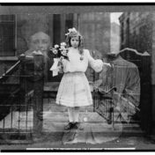Black and white photo of a girl in a white, knee-length dress holding a bouquet of flowers and standing in an open metal gate. The photo is double-exposed so images of two girls and two adult women are faintly superimposed.