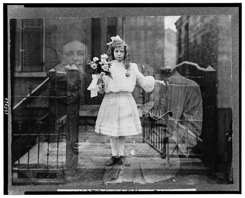 Black and white photo of a girl in a white, knee-length dress holding a bouquet of flowers and standing in an open metal gate. The photo is double-exposed so images of two babies and two adult women are faintly superimposed.