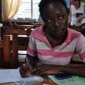 Photograph of a young Black girl. She is wearing a collared shirt with pink stripes and using a pen to write in a notebook.