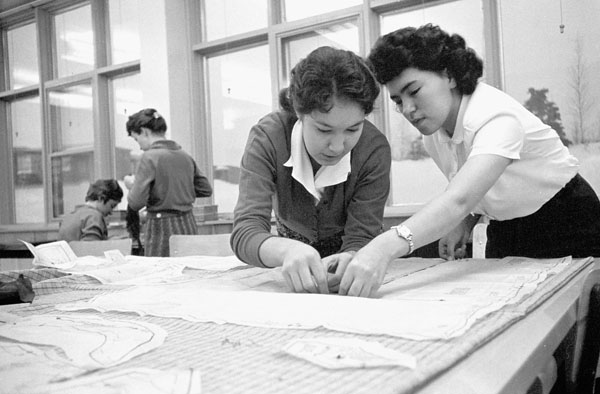 Black and white photo of two teenage girls bent over a table, working with fabric.