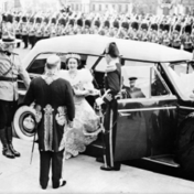 Black-and-white photograph of Queen Elizabeth stepping out of an automobile wearing a long dress and tiara. A man in a tail coat, wearing a sword, is greeting her while a Mountie in uniform salutes.