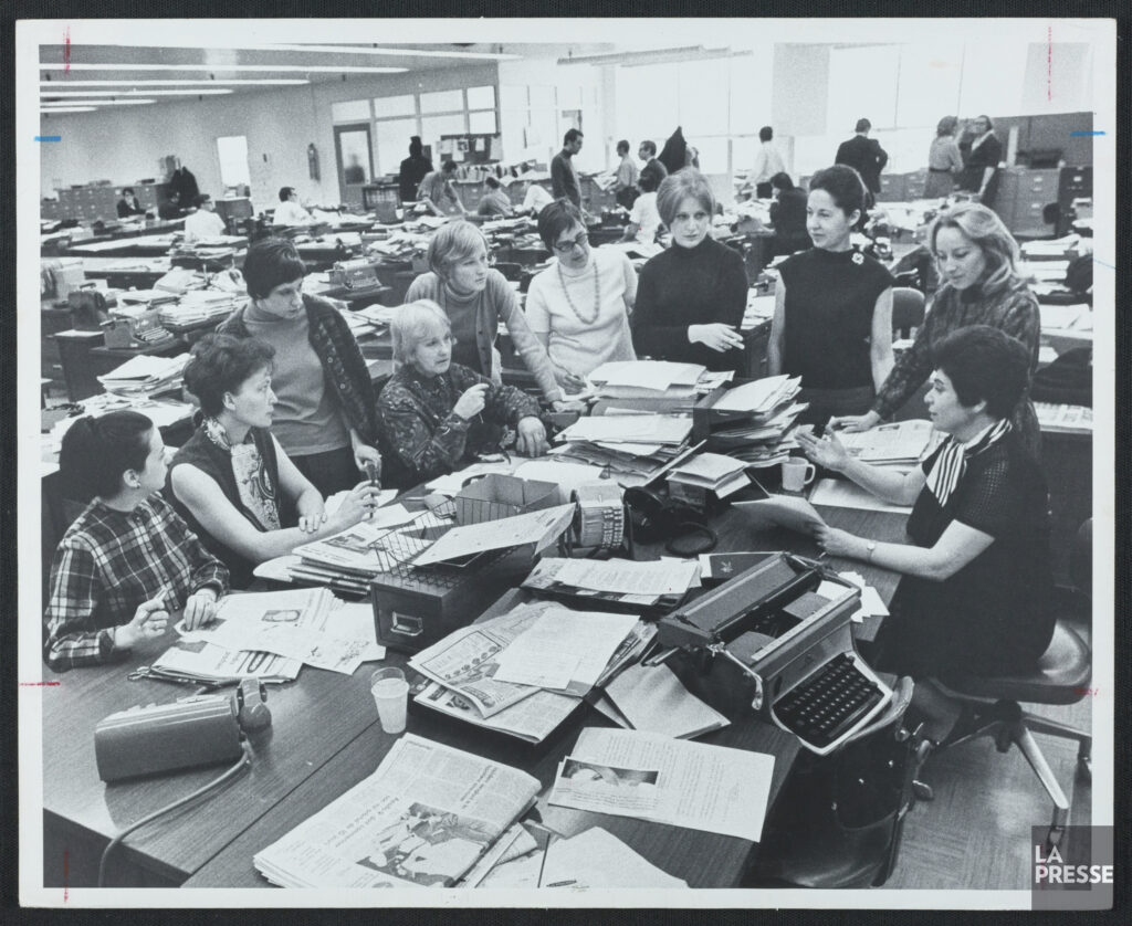 A group of women editors and journalists meet in a busy newsroom.
