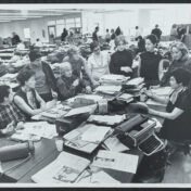 A group of women editors and journalists meet in a busy newsroom.