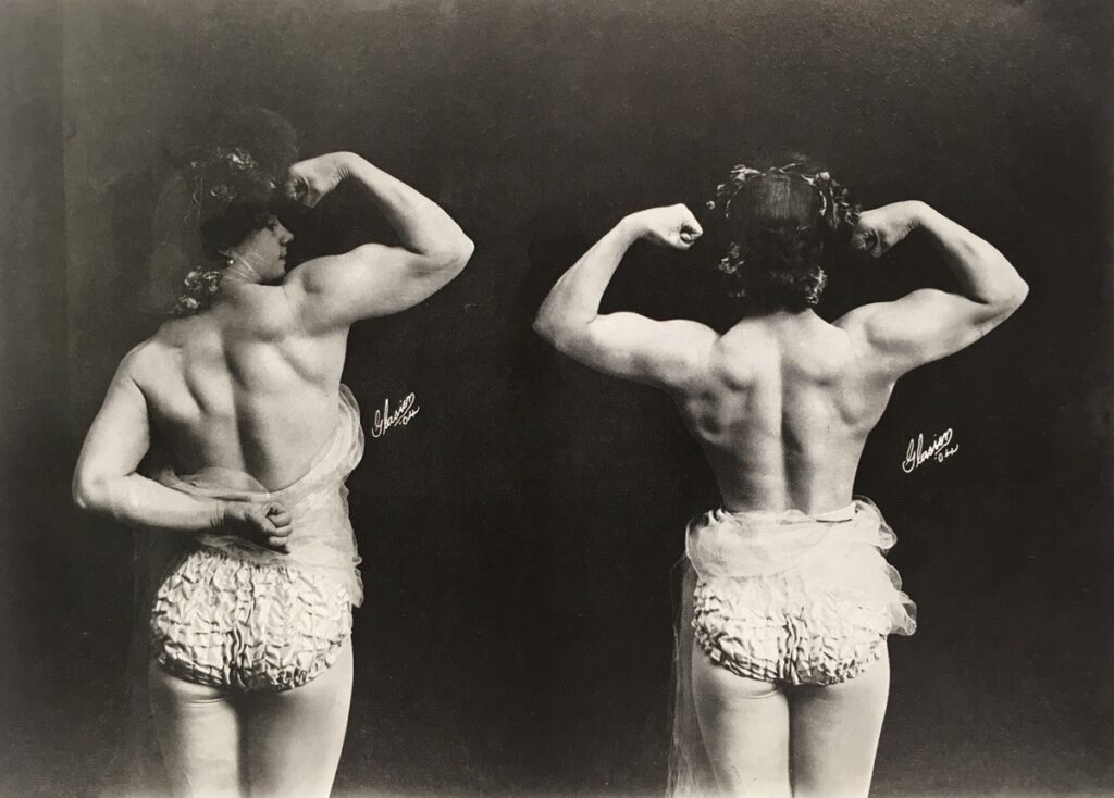 Two black-and-white photos of a woman, viewed from behind. Her hair is curled and in an elaborate updo and she is wearing frilled briefs and wrapped in sheer cloth. She is flexing her muscles to show of the musculature of her back and arms.