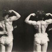 Two black-and-white photos of a woman, viewed from behind. Her hair is curled and in an elaborate updo and she is wearing frilled briefs and wrapped in sheer cloth. She is flexing her muscles to show the musculature of her back and arms.