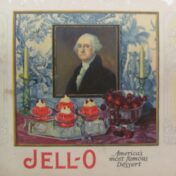 Jell-o advertisement. The tagline is "America's most famous dessert." There is a colour drawing of four dishes of jell-o with dollops of cream and cherries on top, next to a bowl of cherries, between two candlesticks. A portrait of George Washington is behind the table.