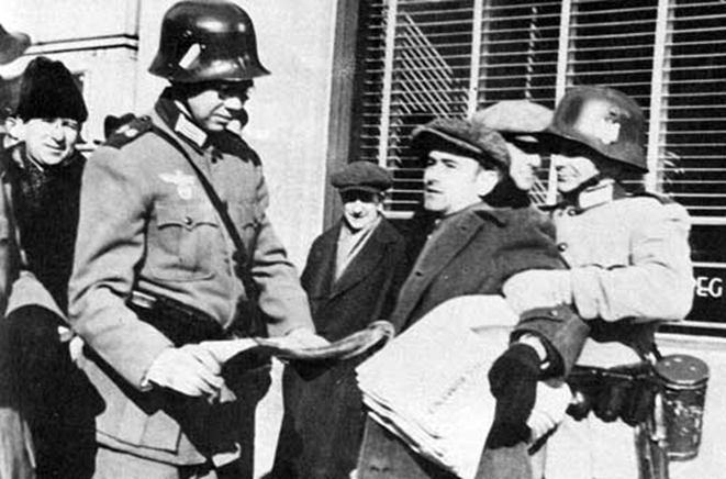 Black and white historical photograph showing a German soldier in a military uniform and helmet reading a document, standing among a group of civilians in coats and caps