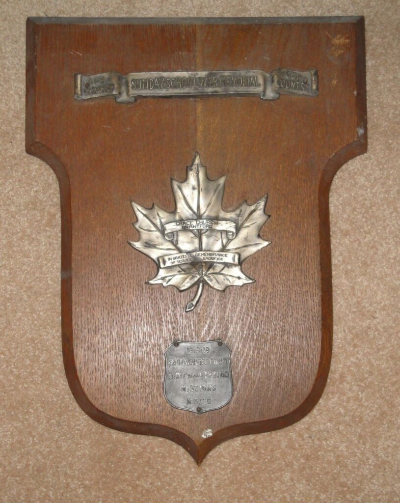 A wooden plaque with metal embellishments. One, a banner at the top, reads "Our Church, Our Country. Sunday School War Memorial." A maple leaf in the centre is embossed with "Grace Church Brantford. In grateful remembrance of service and sacrifice." A shield at the bottom is embossed "1918-19 Endowment Fund Indian and Eskimo Missions MSCC."