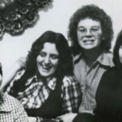 This is a black and white photograph showing four women smiling together in what appears to be a casual, friendly group portrait. They're wearing plaid or checkered shirts, giving the image a relaxed, informal feel. There's decorative artwork visible on the wall behind them - what looks like an ornate floral or scrollwork design. The photo has a vintage quality, likely from the 1970s based on the hairstyles and photographic style. The women appear to be enjoying each other's company, with genuine, warm smiles.