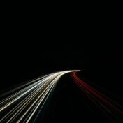 Long exposure photograph of light trails on a highway at night, with white and yellow streaks curving to the left and red streaks on the right against a black background